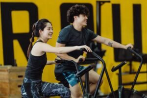 Sporto nauda TAU Asian man and woman enjoying a workout on stationary bikes in a gym, smiling and energetic.